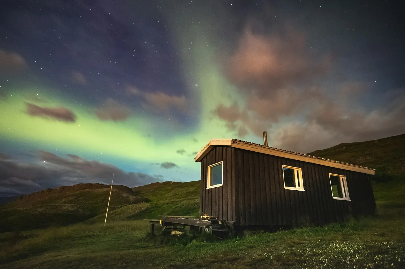 Aurora boreale verde sopra una piccola capanna di legno scuro su un pendio erboso