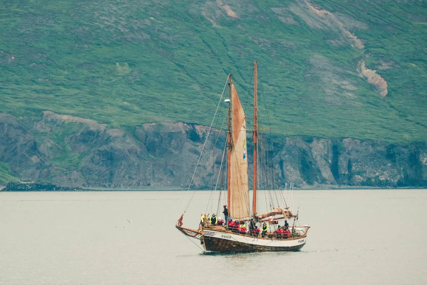 Traditional two-masted wooden schooner with red sails and passengers on calm water in front of a green slope