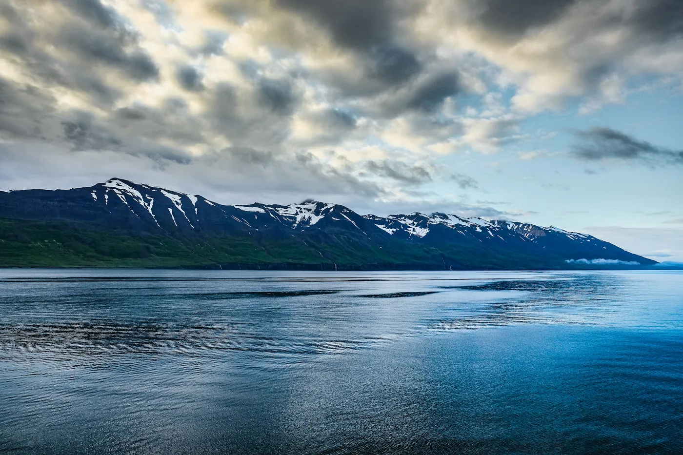 Calm fjord water with a snow-capped mountain ridge under dramatic clouds