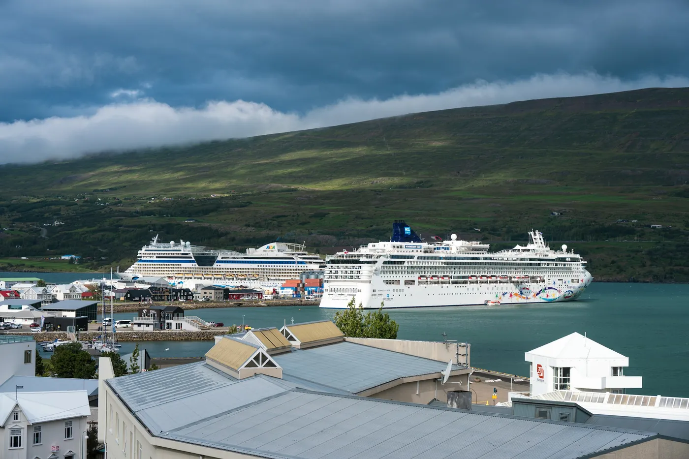 Dos cruceros atracados en el puerto de Akureyri bajo una ladera verde, con tejados en primer plano