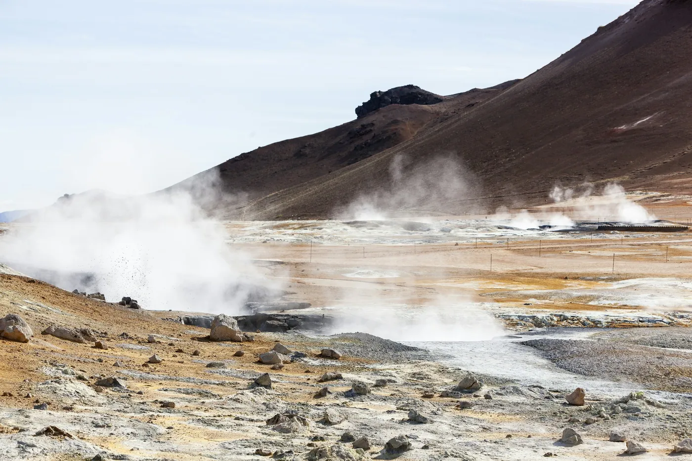 Steaming fumaroles on a bare, ochre geothermal field below a dark hillside