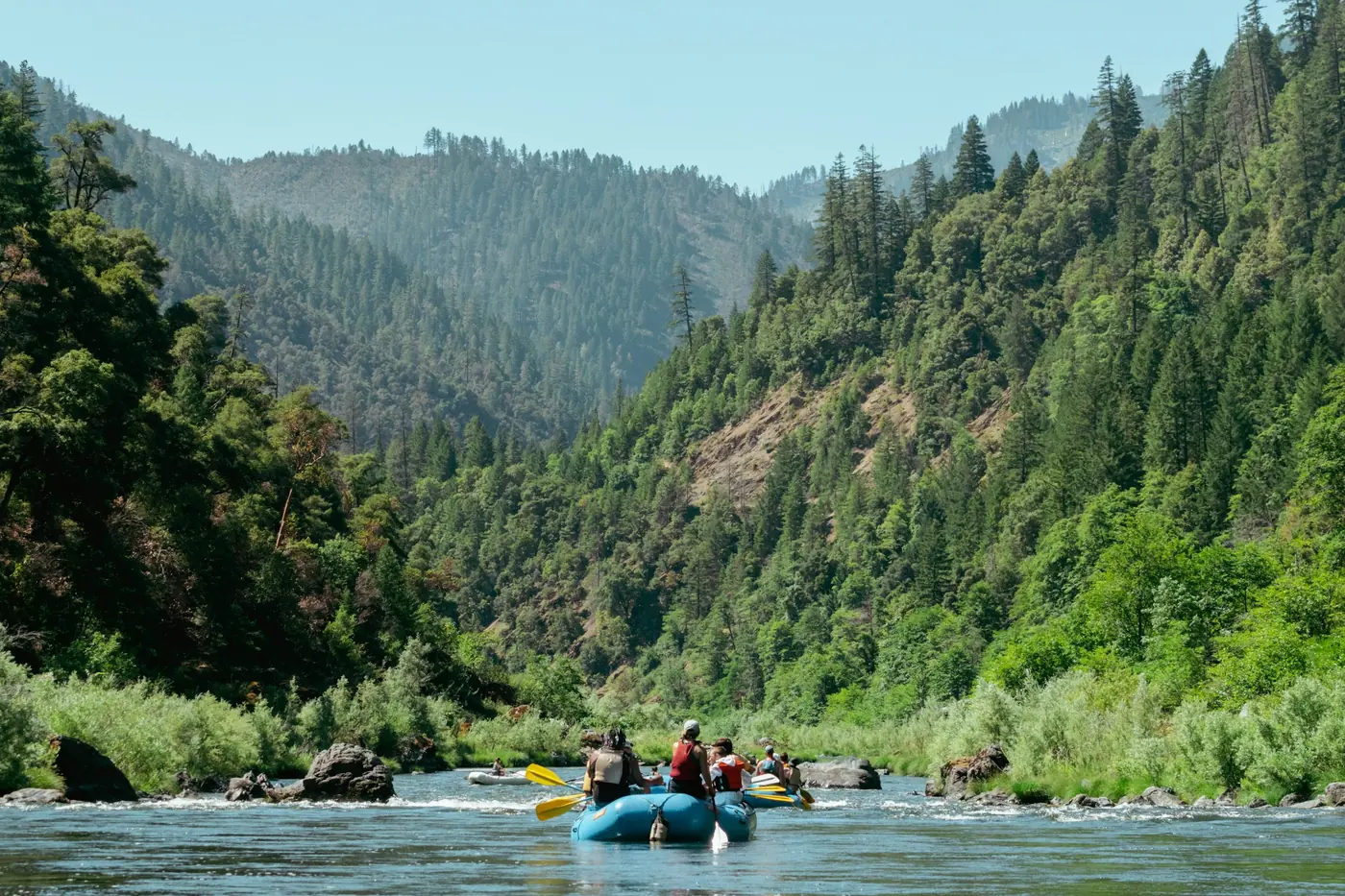 A small group in a blue raft on a clear river between forested mountains