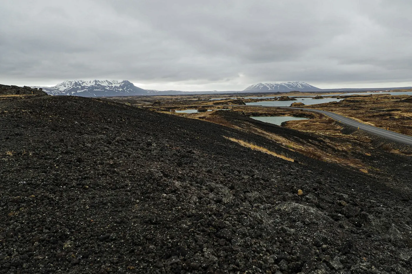 Road curving through a dark lava field towards lakes and distant mountains