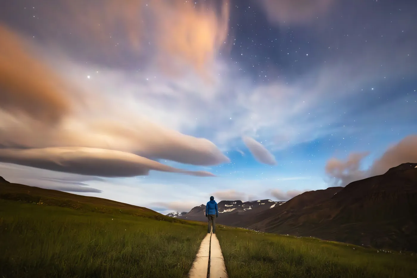 Person in a blue jacket seen from behind on a narrow path through a field, mountains and evening sky